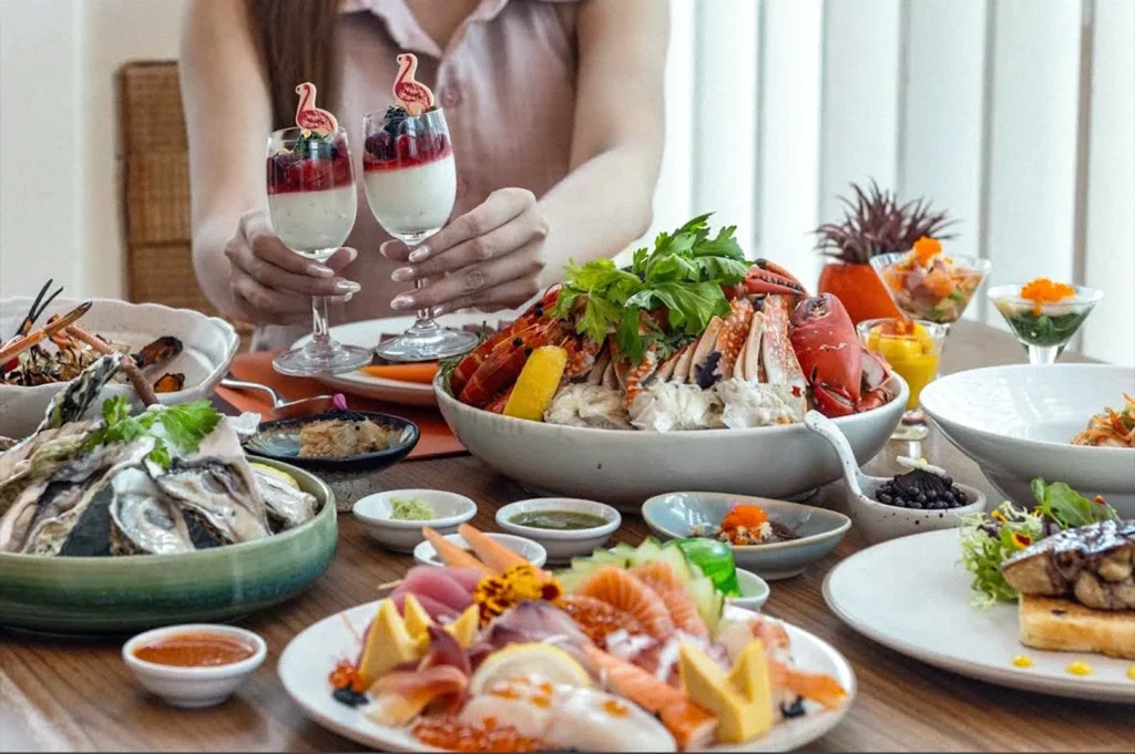 A dining table filled with seafood and international dishes, including lobster, oysters, sushi, and desserts, while a person holds two berry-topped drinks.
