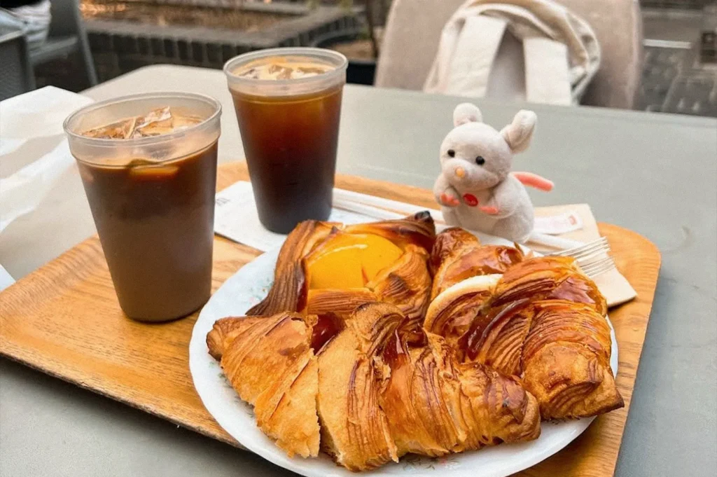 A wooden serving board holds two iced coffee drinks, assorted flaky croissants and pastries, and a small white plush rabbit on a café table.