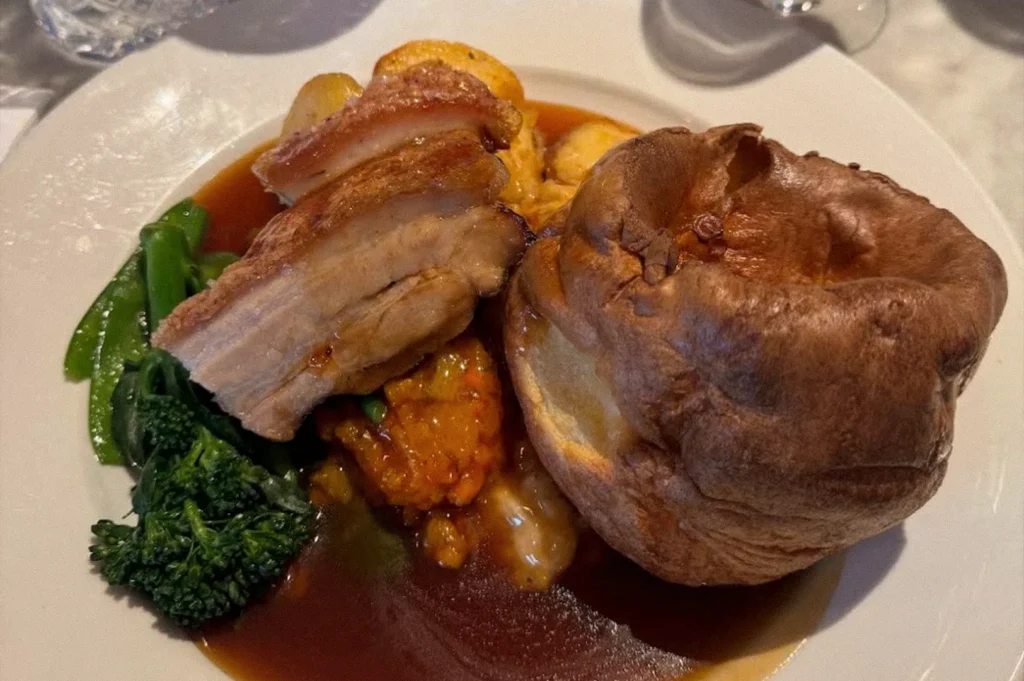 A traditional British roast dinner featuring slices of roast pork with crisp crackling, a Yorkshire pudding, roasted potatoes, green vegetables, and rich brown gravy.