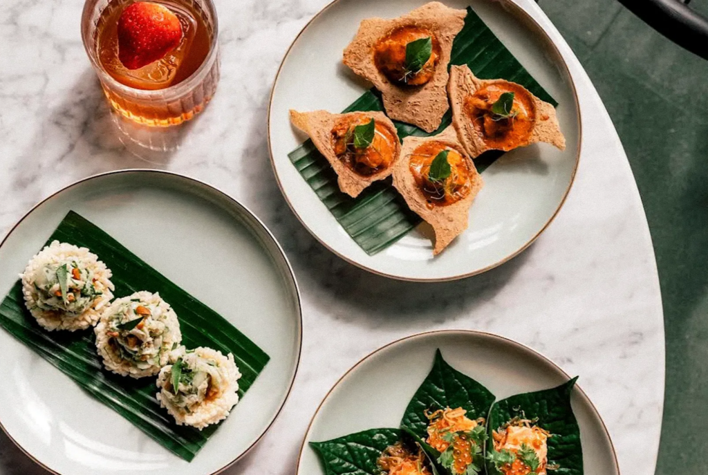 Overhead view of assorted Southeast Asian dishes served on plates lined with banana leaves, including bite-sized savory appetizers and a cocktail with strawberry.