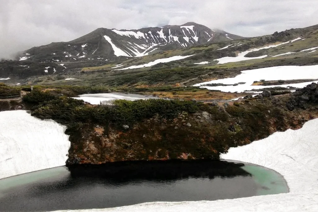 A panoramic view of the Daisetsuzan mountain landscape, featuring rugged ridgelines, scattered snowfields, green alpine vegetation, and a dark reflective pond beneath an overcast sky.
