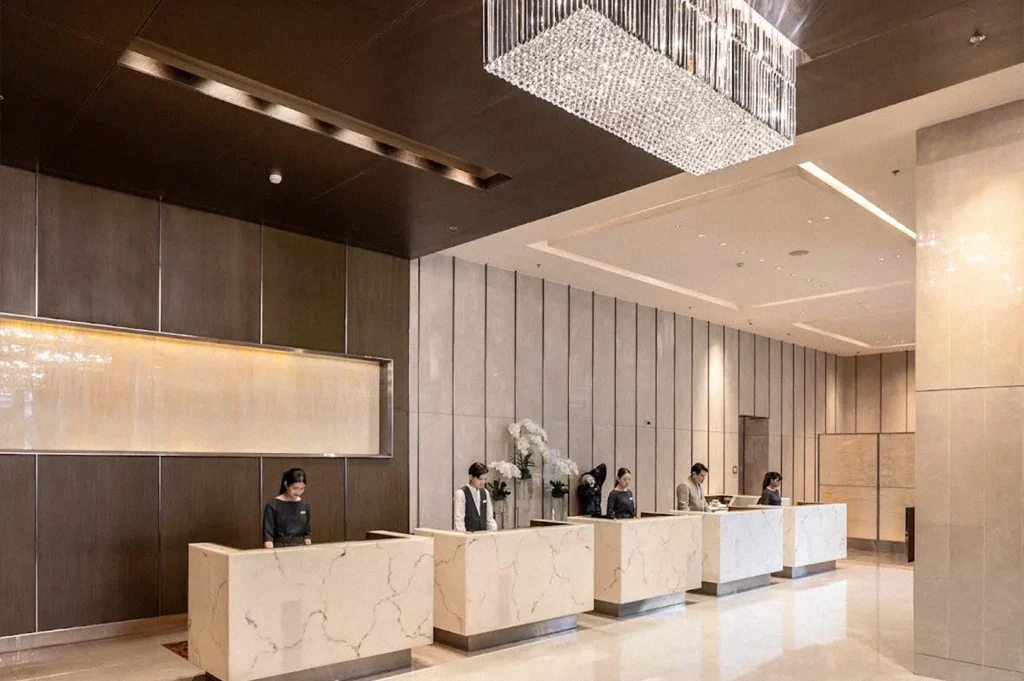 Elegant hotel reception area with several staff members standing behind marble check-in desks beneath a large crystal chandelier.