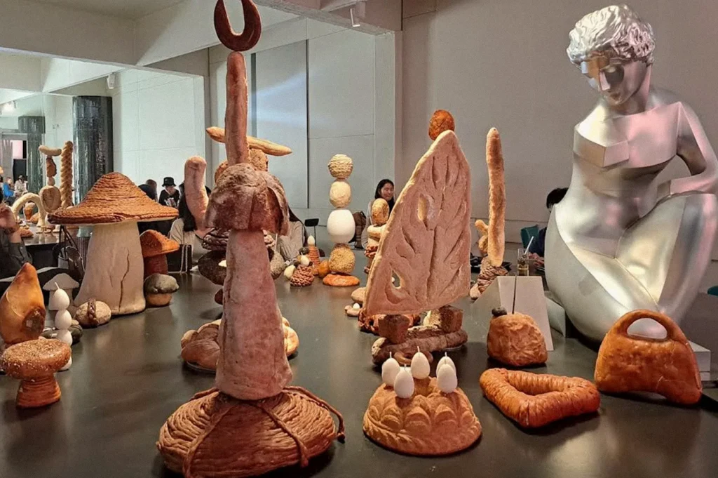 An art features large sculptural forms made of bread, including mushroom-like structures, abstract shapes, and a seated human figure, arranged across a long table in a coffee space.