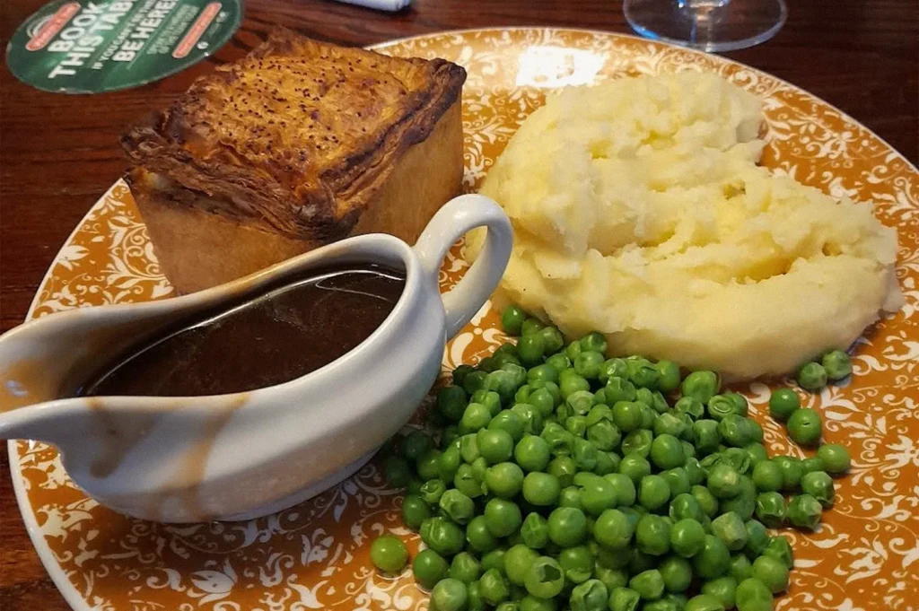 A hearty British pub meal with a golden-crusted savory pie, creamy mashed potatoes, green peas, and brown gravy on a decorative plate.