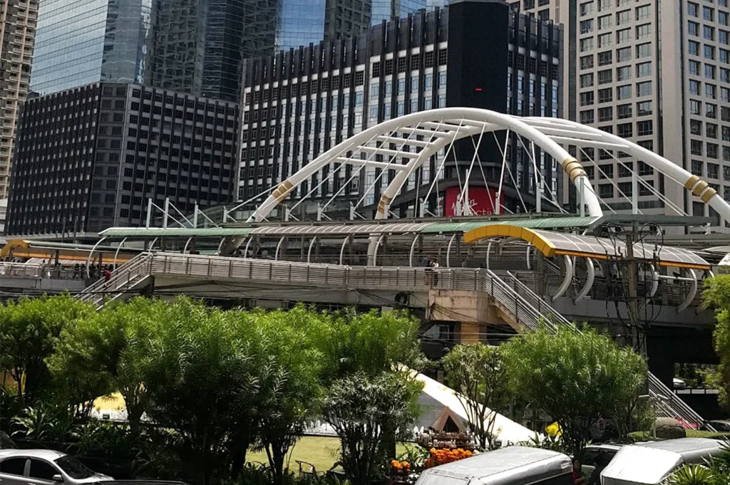 Modern pedestrian bridge with a white arched structure spanning an urban area, surrounded by high-rise buildings, greenery, and parked vehicles.