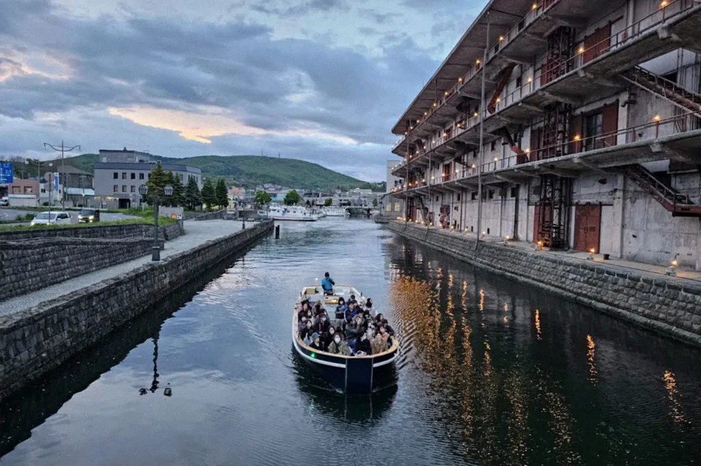 A sightseeing boat carrying passengers along the historic Otaru Canal at dusk, passing stone embankments and a large warehouse building illuminated by warm lights.