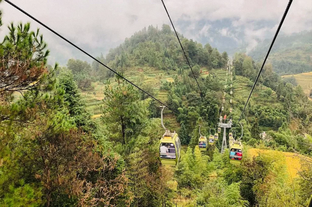 Several cable cars travel along overhead cables above a forested mountain slope, surrounded by green and golden rice terraces beneath a partly cloudy sky.