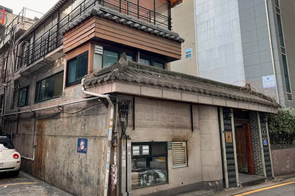 An old Korean-style building with weathered stone walls, wooden accents, and traditional gray tiled roofs stands at a street corner beside taller modern buildings.