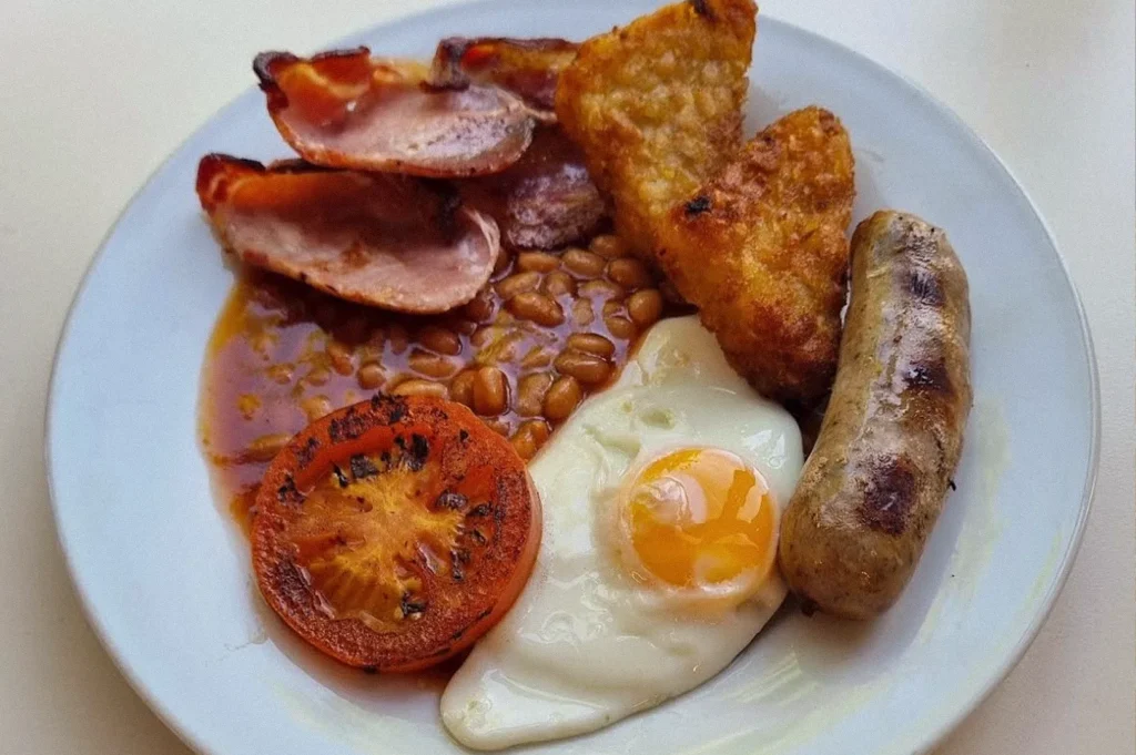 A traditional English breakfast on a white plate featuring grilled sausage, crispy bacon, baked beans, hash browns, a sunny-side-up egg, and a grilled tomato.