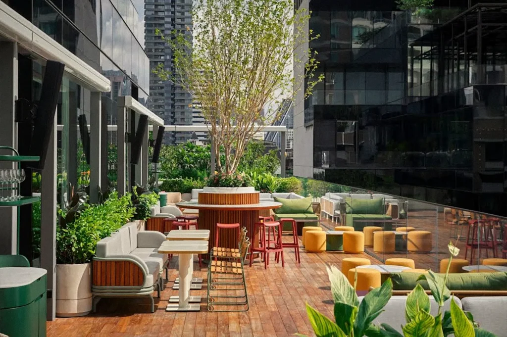 Open-air rooftop terrace with colorful seating, tables, lush plants, and a central tree, surrounded by modern city buildings.
