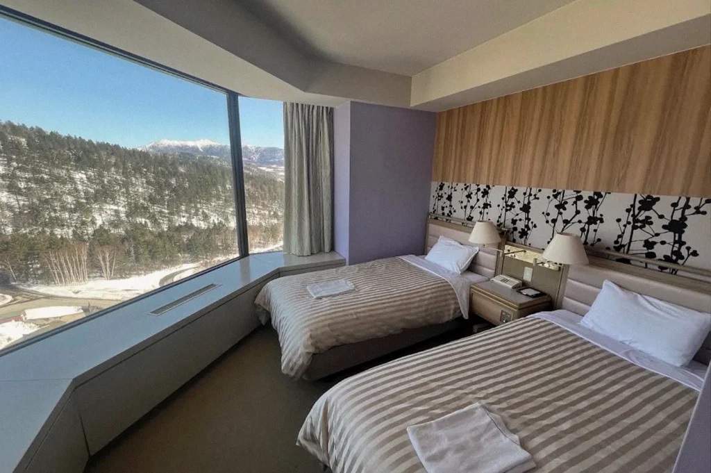 A twin hotel room in Hokkaido with two neatly made beds, wood-paneled walls, and a large panoramic window overlooking snow-covered forests and distant mountain peaks.