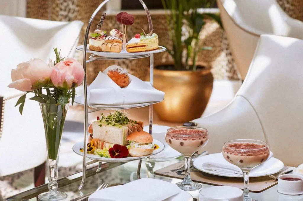 An elegant afternoon tea spread on a tiered stand with assorted pastries, cakes, and finger sandwiches, accompanied by dessert glasses and a vase of pink flowers on a white table.