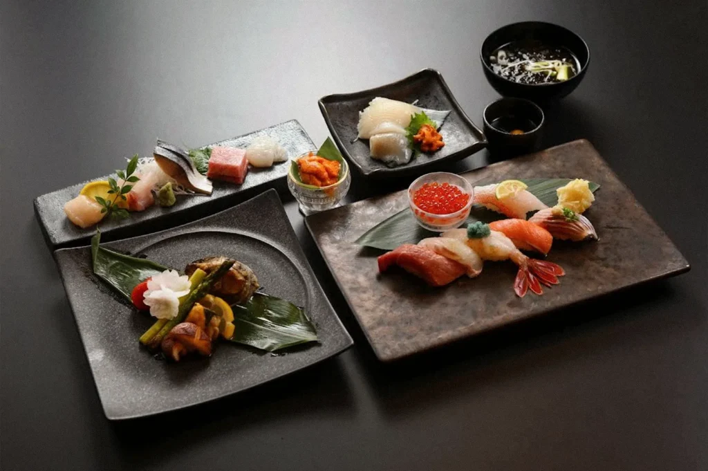 An assortment of Japanese sushi and sashimi arranged on dark ceramic plates, featuring assorted raw fish, shrimp, salmon roe, wasabi, pickled garnishes, and a bowl of soup.