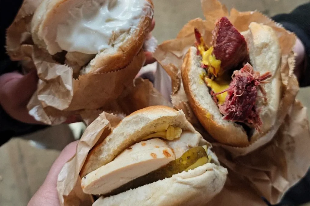 Three handheld bagels in paper wrappers, featuring creamy filling, sliced poultry with pickles, and shredded meat with mustard and cheese.
