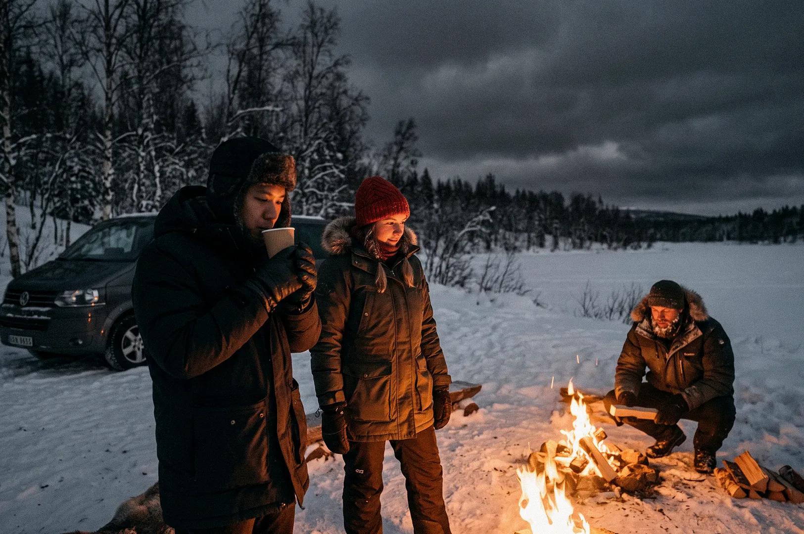 Three people in winter clothing gather around a campfire in a snowy landscape at dusk. One person drinks from a cup while another crouches beside the fire, with a parked van and snow-covered trees in the background.