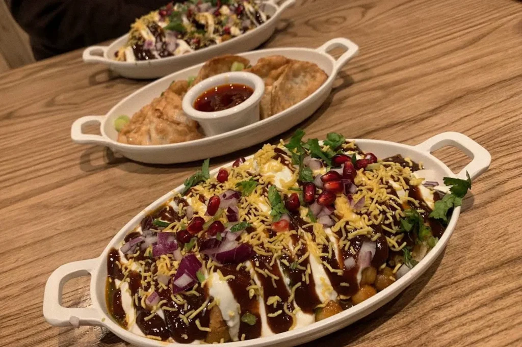 An Indian chaat dish topped with yogurt, dark chutney, crispy sev, pomegranate seeds, chopped red onion, and fresh herbs, with fried dumplings and dipping sauce in the background.