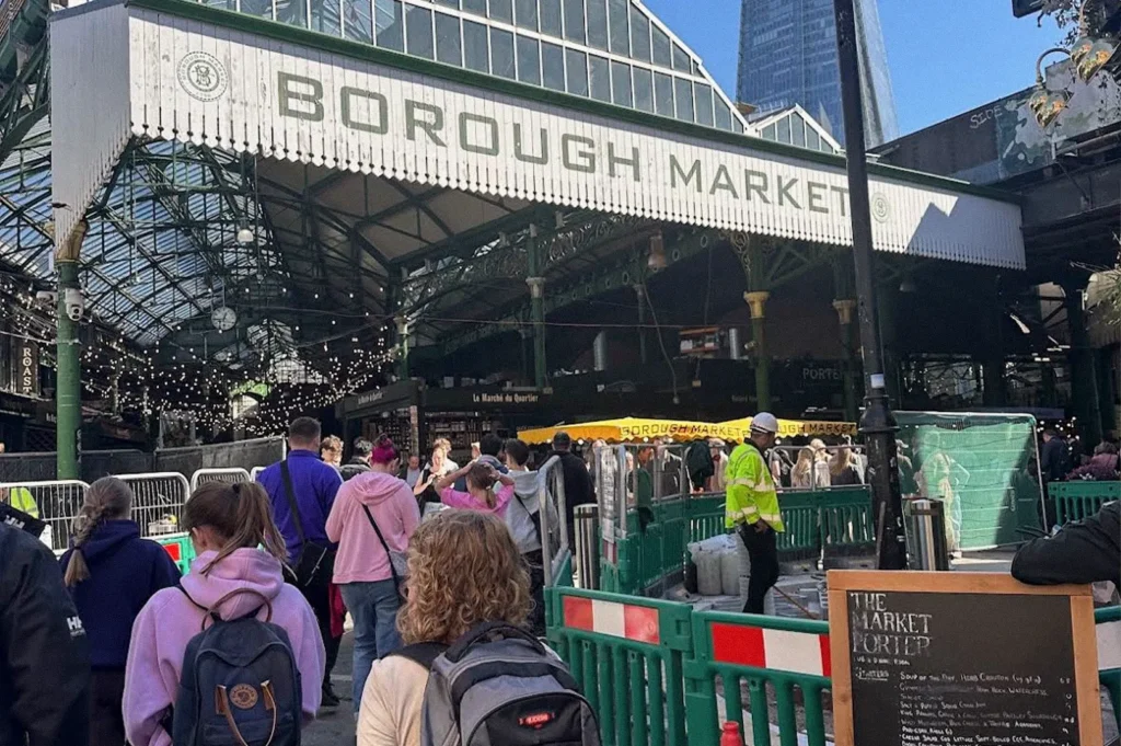The entrance to Borough Market in London, with its large white sign above the covered market and crowds of visitors walking between green barriers beneath the glass-and-steel roof.