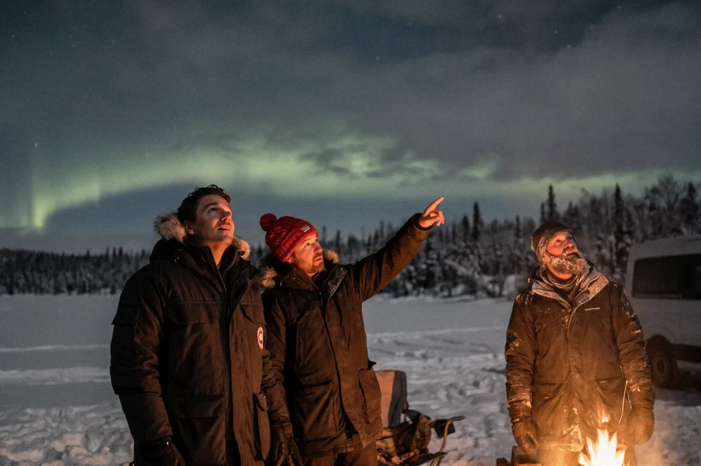 Three people in heavy winter jackets stand beside a campfire in a snow-covered landscape, gazing at the green northern lights illuminating the night sky as one person points toward the aurora.
