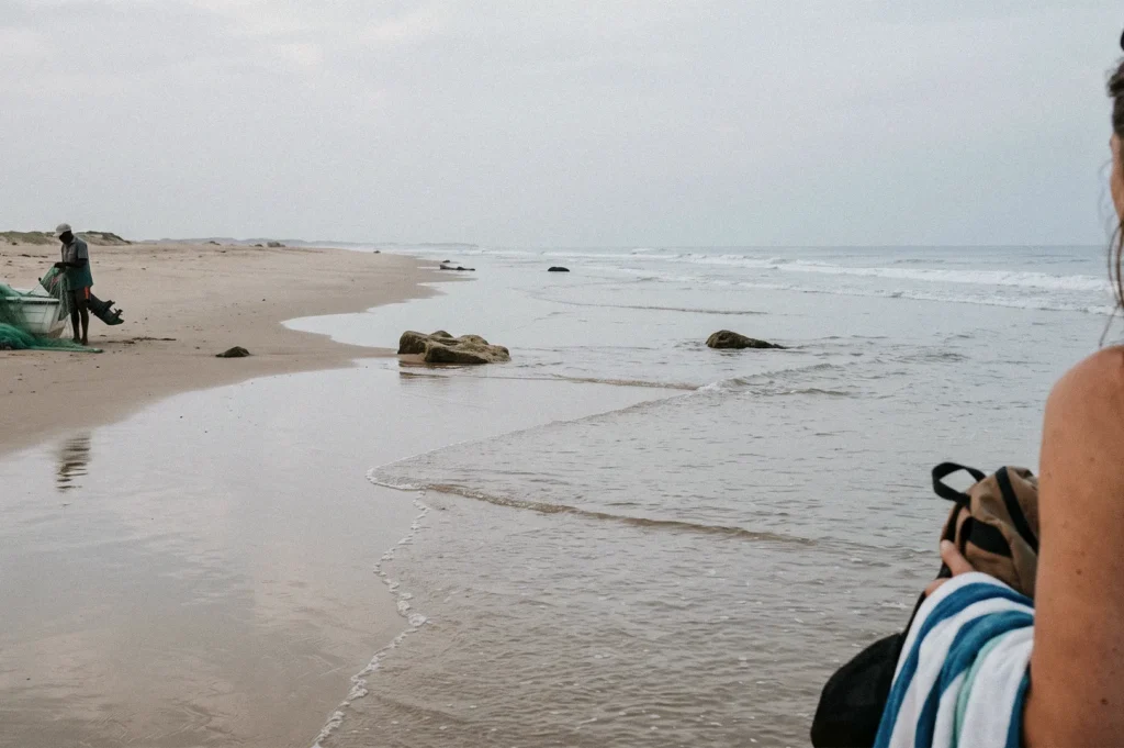 A wide view of a sandy beach with gentle waves washing ashore. A fisherman stands beside his boat and fishing net in the distance, while a female traveler holding a backpack and striped towel is partially visible in the foreground.